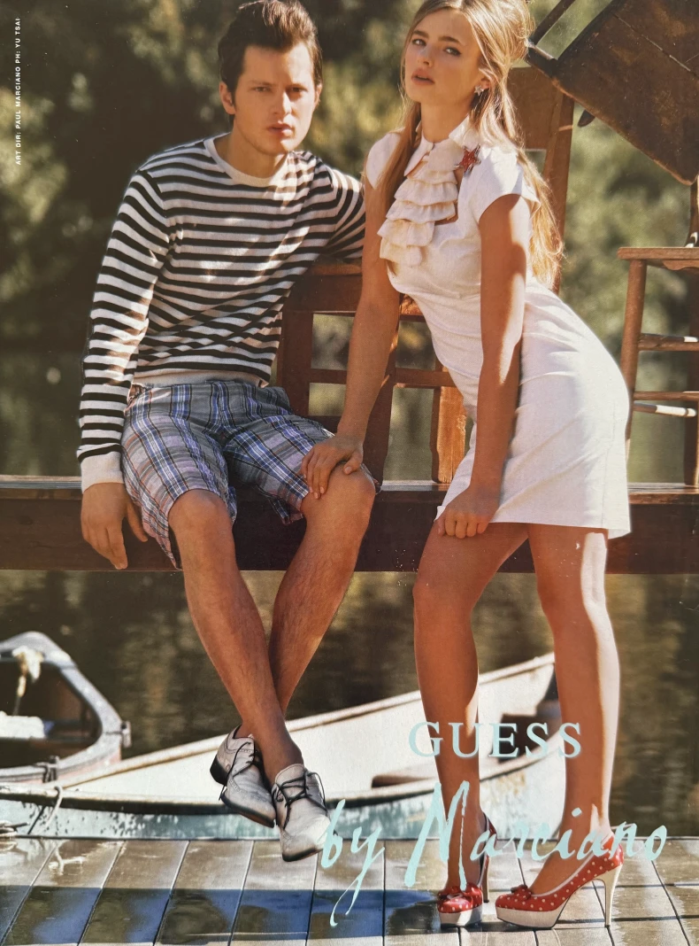 May Gil advertising campaign for Guess by Marciano standing on a jetty, leaning against the railing with one hand on a seated model